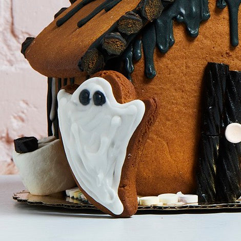 White royal icing ghost cookie leaning against a gingerbread haunted house