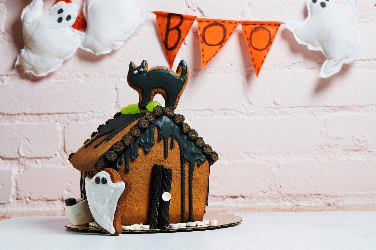 Gingerbread haunted house with black icing drips, a licorice door, a black cat cookie on the roof and a white ghost cookie by the door, in front of a BOO banner